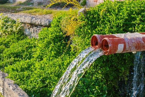 Two weathered orange plastic pipes pour a steady stream of water over a stacked pile of grey stones, creating a small makeshift waterfall surrounded by lush green plants and aged brick walls