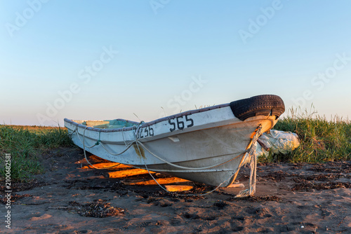 An old white fishing boat rests on dark sandy ground, secured with ropes and a black tire bumper, with a plastic bag nearby, surrounded by beach grass under a clear evening sky