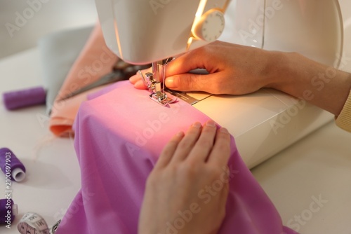 Woman sewing fabric with professional machine at white table in workshop, closeup