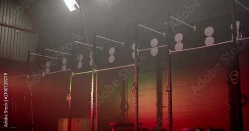 Camera tilting down is revealing full-height warehouse gym rig with climbing rope, boxes, red glow