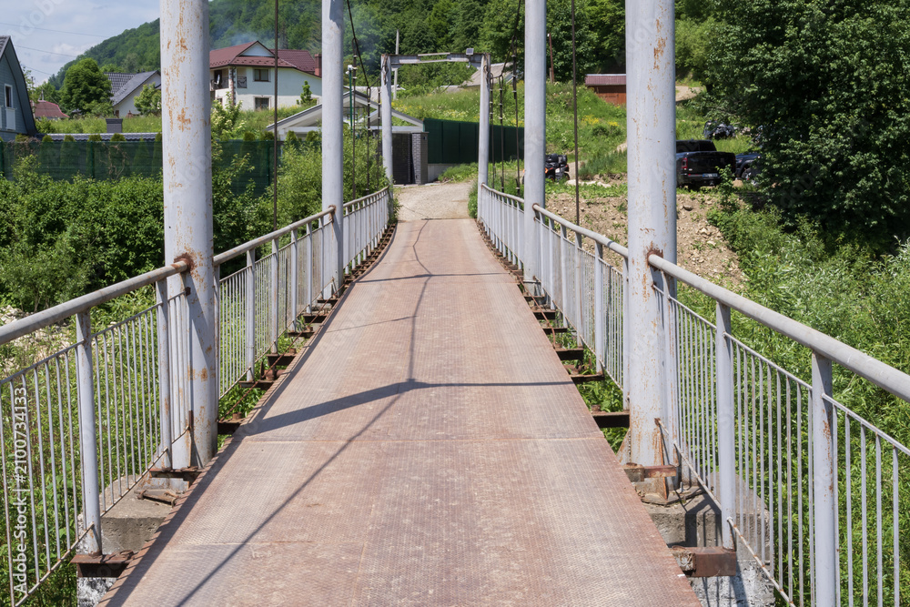 Obraz premium Metal pedestrian suspension bridge in a mountain village with lush green hills. Scenic countryside infrastructure for travel, tourism and transportation concepts.