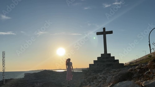 Woman running toward a cross overlooking the ocean at sunset - Christian faith and spiritual journey concept with coastal landscape