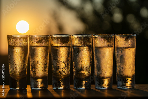 Row of cold shot glasses of vodka or other alcoholic beverages at sunset, closeup. Six chilled alcohol shot glasses filled with clear liquid and covered in condensation drops on golden sunset sky