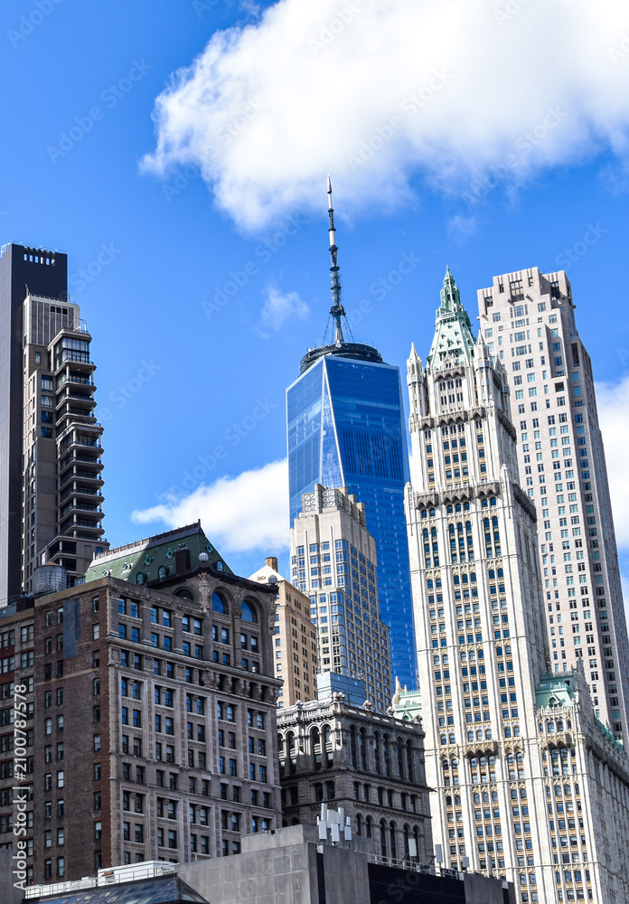 Fototapeta premium Modern skyscrapers of Lower Manhattan including One World Trade Center rise above historic buildings beneath a bright blue sky with scattered clouds in New York City, USA