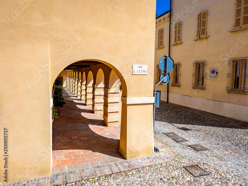 Sabbioneta, Italy, presenting a series of arches and columns along a historical arcade, casting long shadows on the brick floor, connecting to a cobblestone street with traditional buildings
