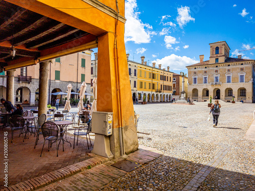 People enjoying outdoor cafes and walking across the cobblestone square bordered by ancient Renaissance buildings under a blue sky in the UNESCO World Heritage site of Sabbioneta, Italy