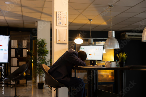фотографія African American man takes an accidental nap while sleeping at desk, weary from overtime and workplace challenges as person struggling with sleep deprivation and burnout, facing intense workload