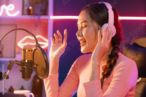 Fotografie Host channel of young beautiful Asian broadcaster listening the music on social media with listeners, wearing pastel headphones, using microphone for singing at neon light indoor studio