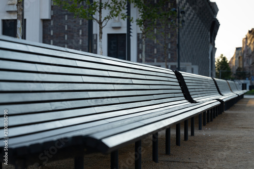 Long row of modern wooden benches with metal frames positioned in a public space, surrounded by trees and contemporary architecture in the background
