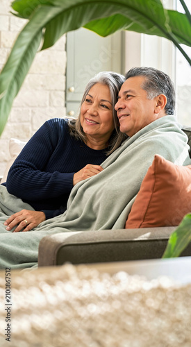 Mature Latina couple sitting on sofa wrapped in a sage green blanket. Happy middle aged Hispanic husband and wife relaxing at home in bright daylight. Retirement lifestyle concept
