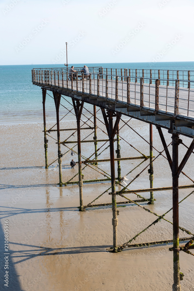 Fototapeta premium Bognor Regis West Sussex England UK. 14.07.2026. Two individuals relax on a pier structure with a weathered appearance, while another person is visible on the sandy beach below