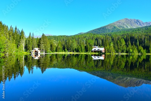Shore of Prebersee lake with cottages and a coniferous forest above and a mirror reflection in the water in Lungau, Niedere Tauern, Austria