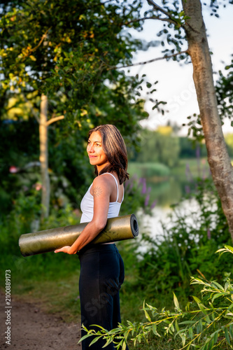 A young Hispanic woman carries a rolled yoga mat along a park path by the lake at golden hour on her way to an outdoor yoga session, embracing an active and mindful summer lifestyle in the fresh air.