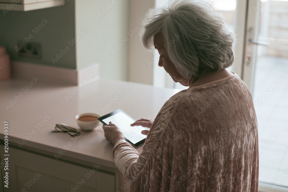 Obraz premium Over-the-shoulder view of woman with tablet at pale kitchen counter