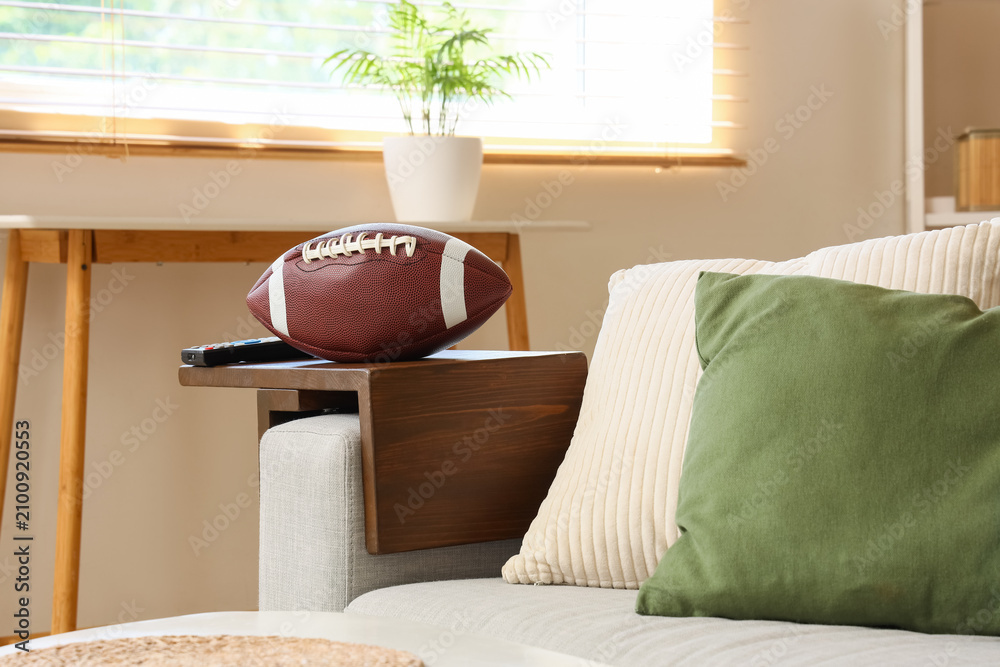 Obraz premium American football and remote control on a wooden sofa armrest tray in a cozy living room setting, the cozy living room features a light-colored sofa with beige and green cushions, a window
