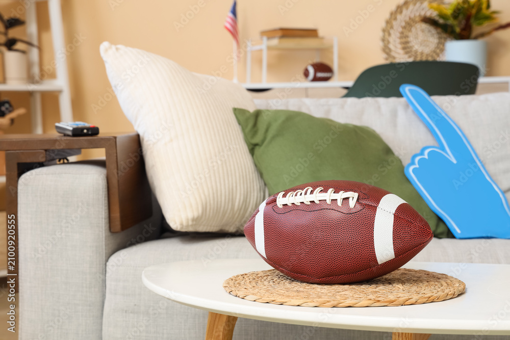 Obraz premium American football on a coffee table in a living room with a foam finger and flag, game day concept, a foam finger and an American flag are visible on the sofa