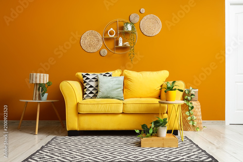 Bild auf Leinwand Comfortable yellow sofa and stylish tables with houseplants near orange wall in