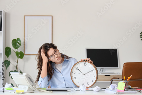Tired young businesswoman with clock at her workspace in office. Deadline con...