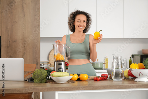 Sporty mature woman with bell pepper and blender making smoothie in kitchen