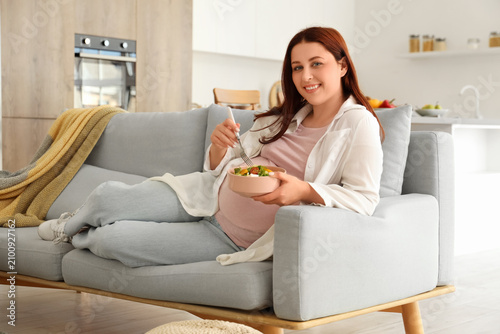 Young pregnant woman with fresh vegetable salad lying on sofa at home