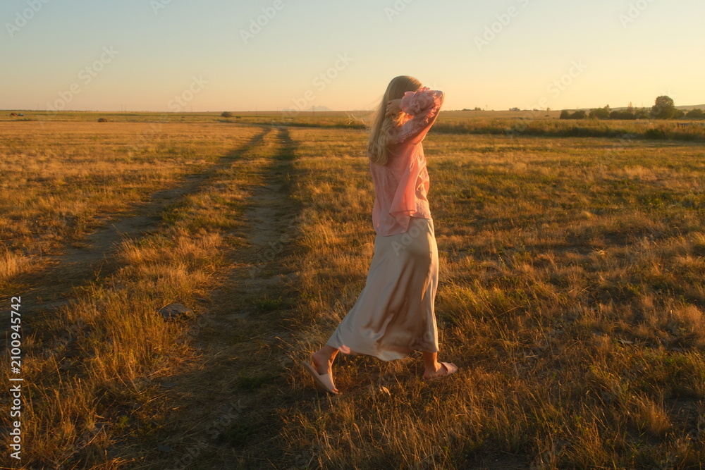 Obraz premium Graceful blond woman in a silk skirt walking along a scenic dirt road in the steppe of East Kazakhstan at sunset. Elegant romantic fashion photography, back view.