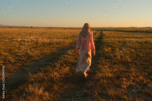 Fotografie Graceful blond woman in a silk skirt walking along a scenic dirt road in the steppe of East Kazakhstan at sunset