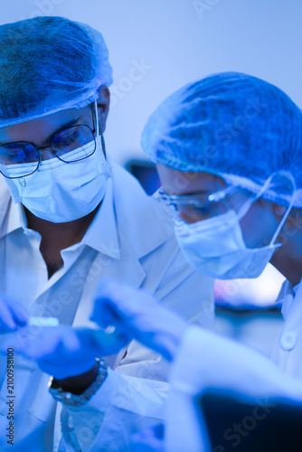 Fotografie Laboratory colleagues work at a microscope in the blue-lit lab in a dramatic blue-lit laboratory while wearing white lab coats and blue protective gloves and modern scientific innovation