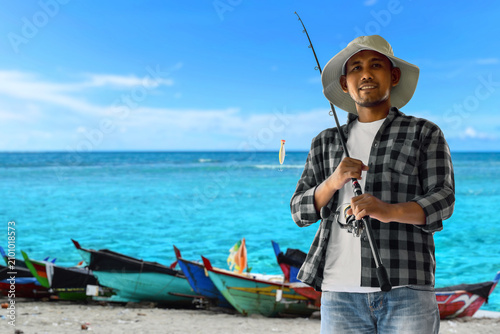 Portrait of man smiling with fishing rod and boats on beach under blue sky