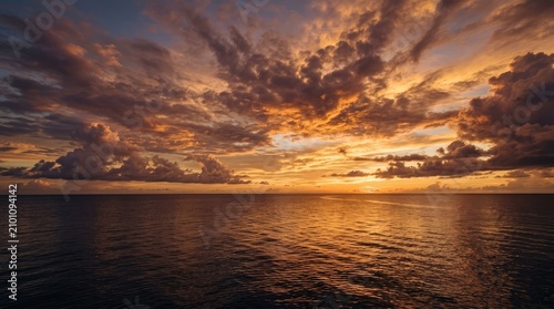 The image captures a breathtaking sunset over the ocean. Vivid orange and golden clouds dominate the sky, reflecting brilliantly on the calm, dark blue water below.
