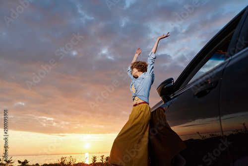 Fotografía A woman stands beside a car at sunset, arms raised in celebration, wearing a denim shirt and flowing skirt
