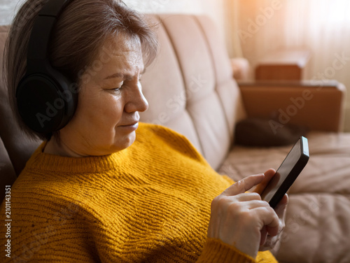 Senior woman relaxing on a sofa, wearing headphones and looking at her smartphone while enjoying music or an audiobook, demonstrating technology use and leisure activity in retirement