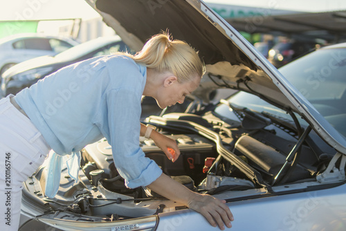 Woman bending over an open car hood, looking into the engine compartment, dealing with a vehicle breakdown and an unforeseen roadside problem