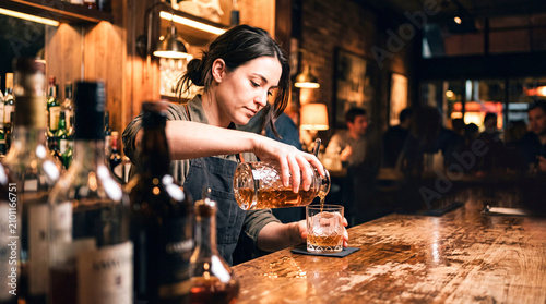 Bartender Pours Whiskey: A focused bartender expertly pours a rich amber whiskey into a glass on a wooden bar, creating a warm and inviting atmosphere.