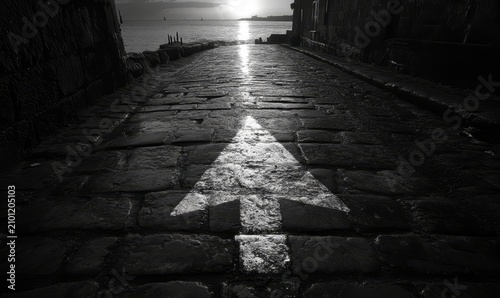 A straight road with an arrow painted on it, leading towards the horizon The sun is setting in the background casting long shadows over cobblestones and asphalt, Generative AI