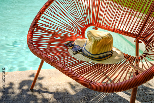 A sunny scene by the pool. Classic summer accessories like a straw hat and dark sunglasses with striking frames on an Acapulco-style bench