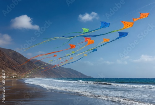 Colorful kites flying over a beach with ocean waves and mountains.