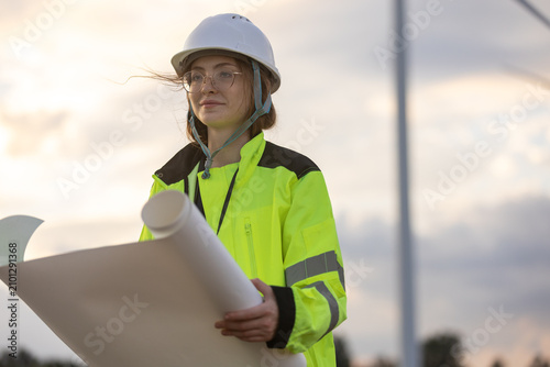 Confident female engineer wearing white hardhat and yellow high visibility jacket holding blueprint outdoors looking focused planning renewable energy project