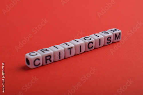 The word criticism spelled with alphabet cubes on a red background, symbolizing evaluation, judgment, feedback, opinion, communication, conflict, and constructive or negative review.