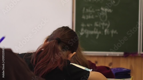 A student writing in a notebook during a classroom lecture, observing math problems on the chalkboard to capture key concepts in an academic setting focused on active learning today