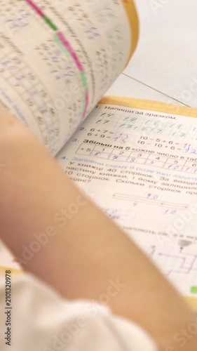 A schoolchild solving math exercises in a workbook with a blue pen on a bright desk, depicting the focused learning process during an early primary education session. Vertical