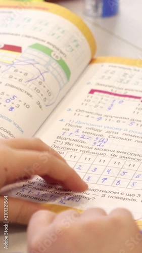 A student solves mathematics homework in an open workbook at a desk, capturing the process of learning arithmetic and basic geometry concepts in a bright classroom setting. Vertical