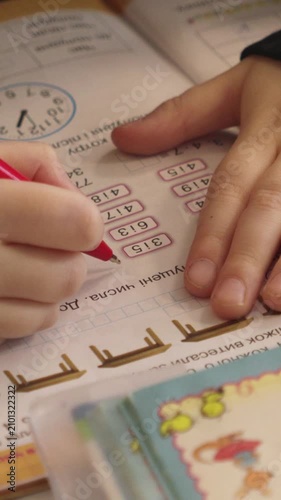 A student hand holds a red pen and writes numbers in an open workbook, illustrating the academic process of learning mathematics through practice in a classroom setting. Vertical
