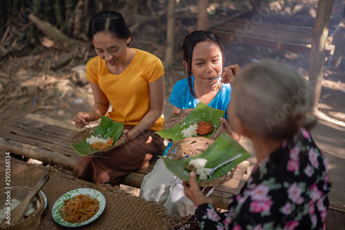Two women and young girl eating homemade food from banana leaf plates on woven baskets beside rustic bamboo seating, capturing family style dining, traditional food culture, and warm outdoor rural
