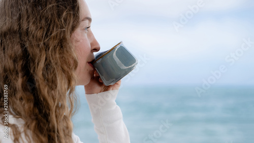 Fotografie Young curly woman enjoying a warm drink while looking at the ocean