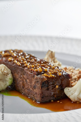 Macro shot of roasted meat glazed with mustard seeds, served with cauliflower and gravy on a white plate. Elegant fine dining close-up.