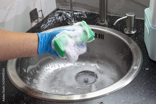 Konstfotografi Close-up of a hand in blue rubber glove holding a soapy foam sponge over a stainless steel kitchen sink