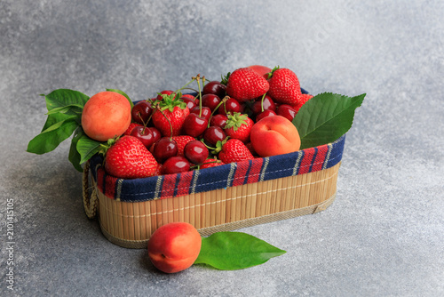 Fresh fruits in a woven basket on a gray surface showcasing a variety of strawberries cherries and apricots arranged neatly