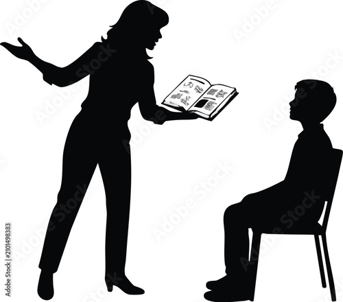 Teacher standing and holding an open book explains a lesson to a student who sits on a chair while listening attentively during an educational session in a classroom.
