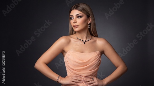 Gorgeous young woman looking aside, wearing luxury black onyx and rose gold jewelry set, statement necklace, drop earrings, matching bracelet, and ring, peach strapless draped chiffon dress, studio po