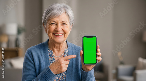 Mobile Advertising, Chroma Key, Smiling senior woman shows mobile phone with green screen pointing with index finger at smartphone Chroma key mock-up